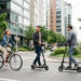 Rue moderne avec des usagers en mobilité urbaine : une femme à vélo, un homme en trottinette et un homme en scooter électrique, en déplacement au milieu des bâtiments contemporains.