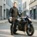 Jeune homme équipé d'une tenue de motard debout devant sa moto, casque à la main, dans une rue urbaine sobre éclairée par la lumière matinale.