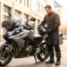 Un homme équipé d'une tenue de motard ajuste la bagagerie d'une moto de tourisme dans une rue urbaine lumineuse, avec son casque à la main.