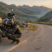 Un motard équipé fixe ses bagages près d'une moto de voyage sur une route panoramique bordée de montagnes, éclairée par une lumière dorée.
