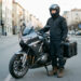 Un homme motard équipé d’un casque, d’une veste de protection et de sacoches se tient près de sa moto, avec une route et une ville en arrière-plan.